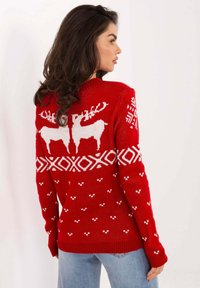 Frau im Profil, die einen roten Weihnachtspullover mit weißen Rentieren und geometrischen Mustern trägt, kombiniert mit hellblauen Jeans.