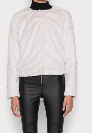 Vrouw draagt een witte, getextureerde jas van namaakbont over een zwarte coltrui en zwarte leren broek met ritssluiting aan de voorkant.