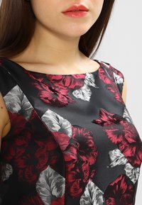 Šaty s květinovým potiskem s velkými červenými a stříbrnými květinami na černém saténovém materiálu. Hranatý výstřih a přiléhavý střih, detaily na ramenech.