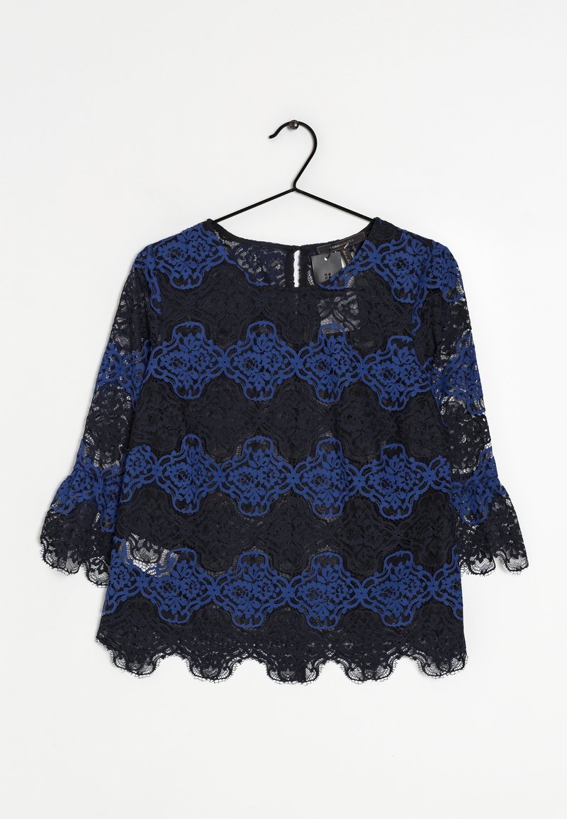 Blouse en dentelle noire et bleue avec motif floral, manches trois-quarts, bords festonnés, suspendue sur un cintre noir sur fond blanc.