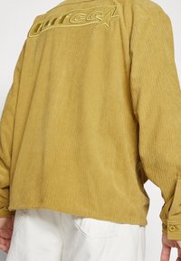 Giacca di velluto a coste gialla con vestibilità rilassata, caratterizzata da un logo ricamato sul retro e polsini con bottoni, abbinata a pantaloni bianchi.