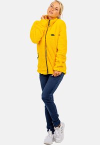 Geel fleecejack met ritssluiting aan de voorkant, twee zijzakken en een ronde logopatch. Draag het over donkere spijkerbroeken en witte sneakers. Textuurstof.