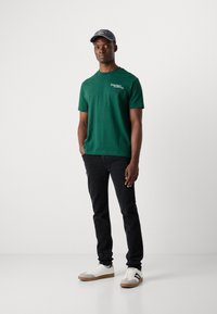 Green cotton t-shirt with white text, paired with black jeans and beige accented sneakers. The model wears a grey cap.