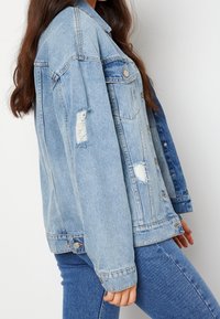 Vaaleansininen denimtakki rentolla istuvuudella, kulutuksilla, suurilla taskuilla ja metallisilla nappeilla, yhdistettynä samansävyisiin kapealahkeisiin farkkuihin.