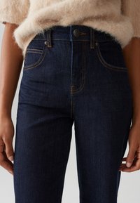 Donkerblauwe denim jeans met een aansluitend ontwerp, uitgerust met koperen stiksels, een riem met knoopsluiting aan de voorkant en een klassieke vijf-pocket stijl.