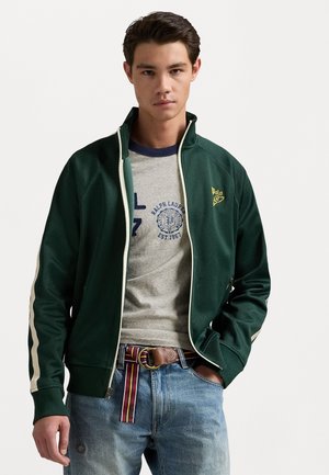 Chaqueta verde con cremallera y rayas crema, llevada sobre una camiseta gris con estampado de logo azul. Vaqueros azules y un cinturón multicolor.