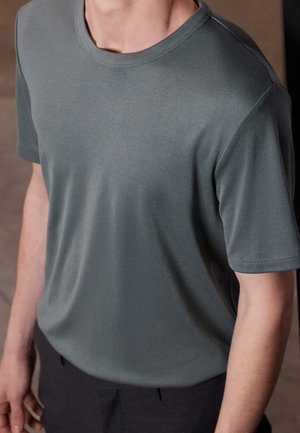 Homme portant un t-shirt gris foncé uni à manches courtes et col rond, et un pantalon noir, visible du cou à la taille.