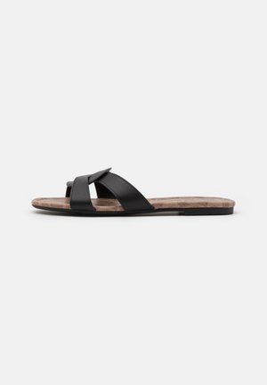 Sandale plate style mule avec larges brides croisées en cuir noir et semelle intérieure marron texturée sur fond blanc.