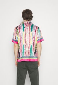Mann mit dem Rücken zur Kamera, der ein buntes, gemustertes Kurzarmhemd mit abstrakten Designs in Pink, Grün, Gelb und Schwarz trägt, kombiniert mit dunklen Hosen.