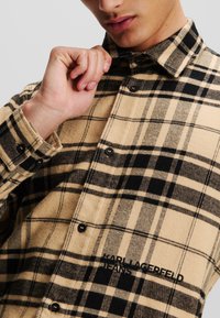 Camisa de franela beige con patrón de cuadros negros, cuello tipo clásico, cierre de botones y bordado "KARL LAGERFELD JEANS" en el pecho.