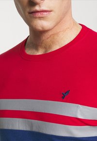 T-shirt rossa con strisce orizzontali grigie e blu, scollatura rotonda e piccolo logo di uccellino ricamato blu sulla parte sinistra del petto.