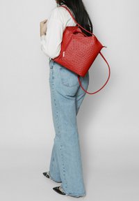 Rode leren pull-string rugzak met een textuurpatroon, voorzien van een zijritssak en een enkele schouderriem. Wijdvallende jeans maken de look compleet.