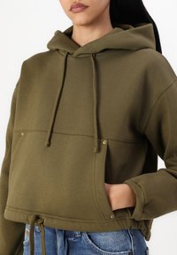 Λαδί πράσινη κοντή ζακέτα με κουκούλα, με κορδόνι, μπροστινή τσέπη και ορειχάλκινες λεπτομέρειες. Κατασκευασμένη από μαλακό, υφασμάτινο υλικό με άνετη εφαρμογή.