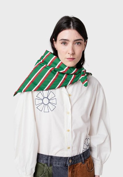 Sciarpa a righe verdi e rosse appoggiata su una camicia bianca con bottoni. La sciarpa ha una trama liscia; la camicia presenta ricami circolari sul petto.