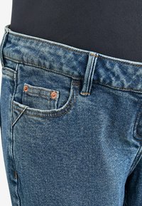 Jeans de mezclilla azul con un corte estándar, que presentan un pequeño bolsillo frontal, remaches de cobre y costuras visibles a lo largo del cinturón y las costuras.