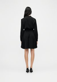 Γυναίκα που στέκεται με την πλάτη προς την κάμερα, φορώντας ένα μαύρο φόρεμα με μακριά μανίκια και μαύρες ψηλοτάκουνες γόβες, μπροστά σε λευκό απλό φόντο.