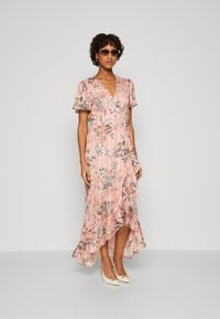 Maxi dress in light pink fabric with floral print, short flutter sleeves, V-neck, ruffled hem, and paired with white pointed toes shoes.