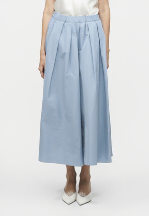 Γυναίκα που φοράει ανοιχτό μπλε παντελόνι φαρδύ και κοντό με πιέτες και ελαστική μέση, σε συνδυασμό με λευκές ψηλοτάκουνες μυτερές γόβες.