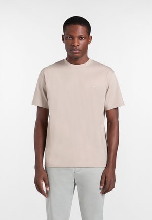 Mężczyzna ubrany w beżowy T-shirt z krótkim rękawem i jasnoszare spodnie stoi na tle jednolitego białego tła.