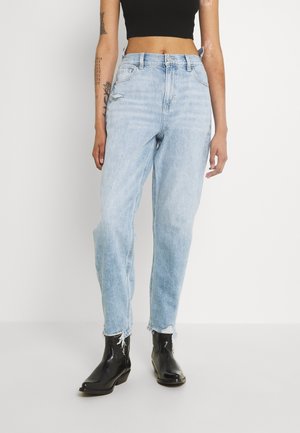 Persoon draagt lichtblauwe versleten jeans met hoge taille, zwart cropped topje en zwarte enkellaarzen, staand tegen een effen witte achtergrond.