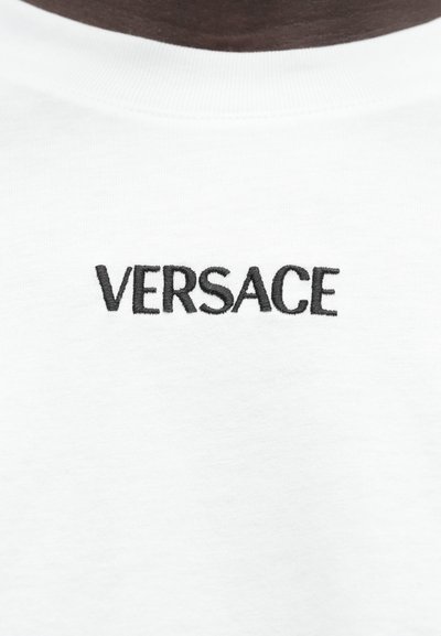 Biała bawełniana koszulka z czarnym haftowanym logo "VERSACE" umieszczonym na środku klatki piersiowej. Miękka w dotyku o regularnym kroju.