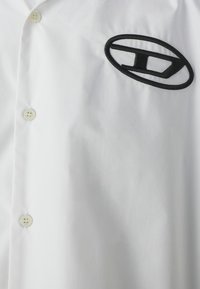 Hvid skjorte med knaplukning og glat overflade. Har et broderet sort logo på venstre side og standard plastknapper.