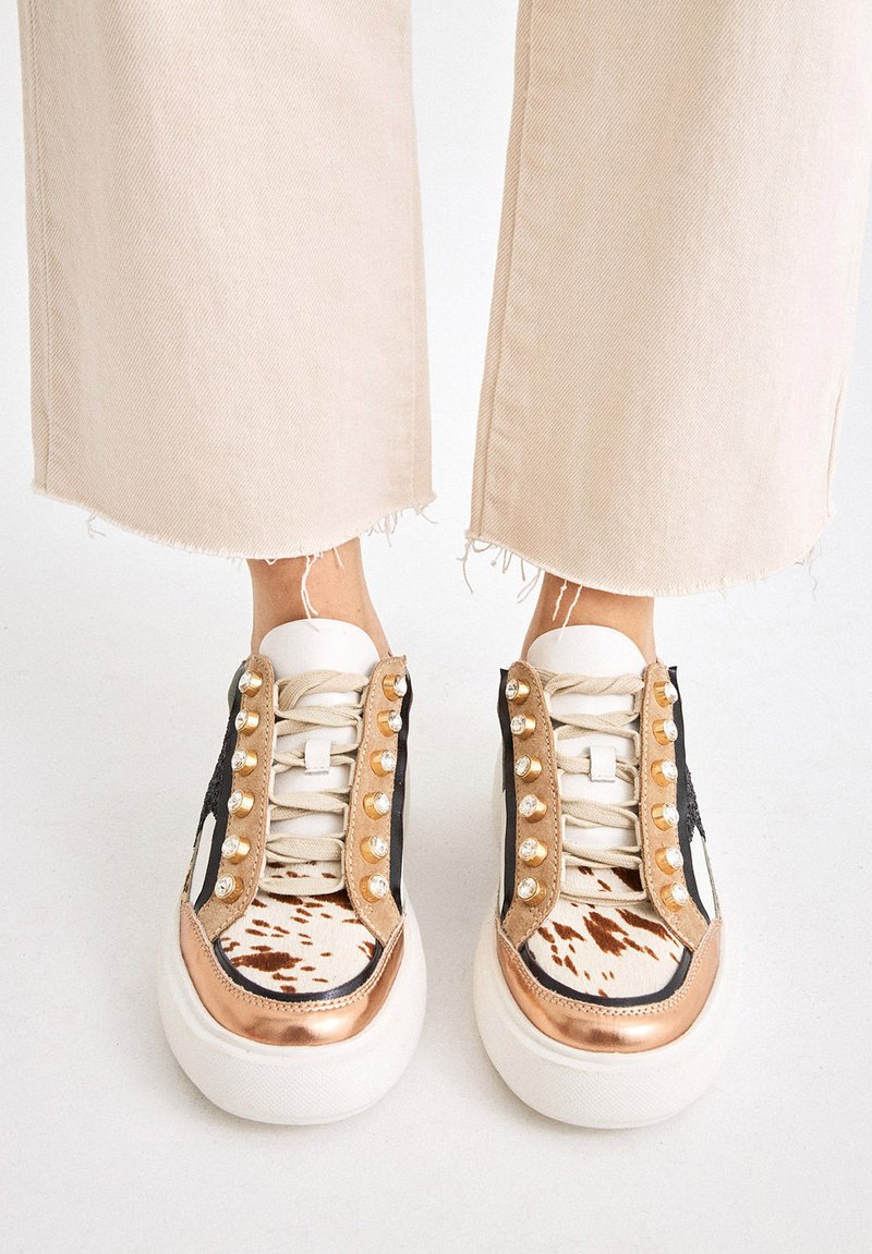Persona con pantalones de pierna ancha beige y zapatillas multicolor con tachuelas doradas, cordones blancos y detalles de estampado animal.