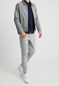 Šedá lehká bunda s límečkem přes námořnickou košili s knoflíky, doplněná o světle šedé vzorované kalhoty a bílé tenisky.