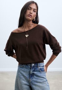 Brauner schulterfreier Pullover mit langen Ärmeln, kombiniert mit hellblauen High-Waist-Jeans. Schmuck umfasst einen herzförmigen Anhänger.