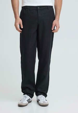 Mann trägt schwarze, gerade geschnittene Hosen und weiße Sneakers mit schwarzen Streifen, steht vor einem schlichten hellen Hintergrund.