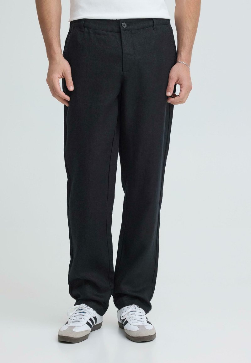 Άνδρας που φοράει μαύρο παντελόνι με ευθείες γραμμές και λευκά αθλητικά παπούτσια με μαύρες ρίγες, στέκεται μπροστά σε μια απλή ανοιχτόχρωμη επιφάνεια.