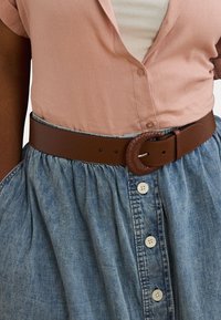 Brun lærbelte med en flette detalj, brukt over et lyst denimskjørt med knapp foran og samlet liv. Rosa skjorte under.