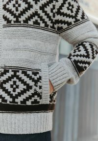 Grå strikket sweater med sorte geometriske mønstre på ærmerne og kanten. Har ribbede manchetter og en høj hals for varme.