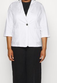 Osoba oblečená v bílém saku na jedno knoflík s třičtvrtinovými rukávy, pod kterým má černý top a černé kalhoty, stojí na jednobarevném pozadí.