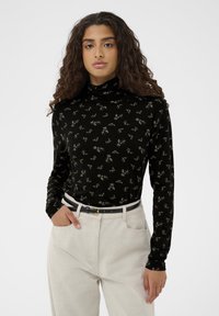 Čierny rolák s drobným kvetinovým vzorom v bielej a zlatej farbe, vyrobený z mäkkého materiálu, v kombinácii so svetlo béžovými nohavicami a čiernym opaskom.