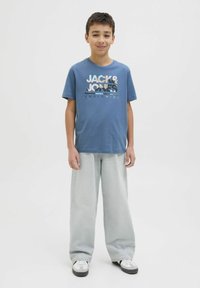 Kék pamut póló, fehér és szürke "JACK & JONES" logóval, világosszürke, bő szárú farmernadrággal és fekete-fehér sportcipőkkel párosítva.