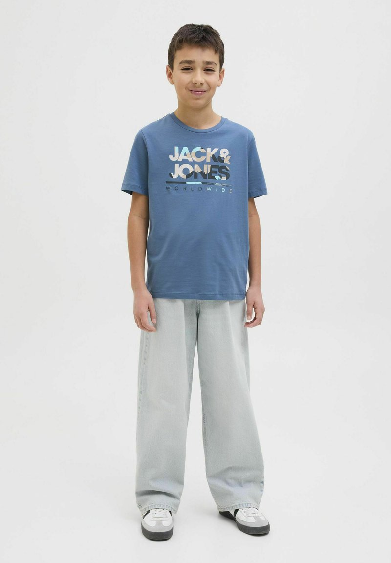 Kék pamut póló, fehér és szürke "JACK & JONES" logóval, világosszürke, bő szárú farmernadrággal és fekete-fehér sportcipőkkel párosítva.
