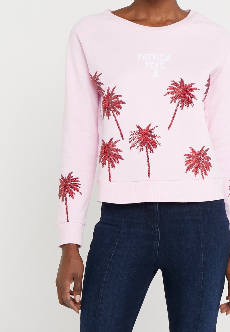 Femme portant un sweat-shirt rose clair avec des motifs de palmiers rouges en sequins et un jean bleu foncé, torse et haut des jambes visibles.