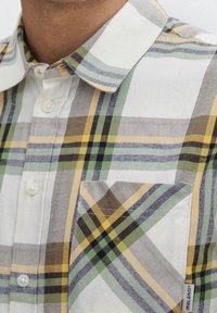 Nahaufnahme eines Mannes, der ein kariertes Hemd in Weiß, Grün, Gelb und Schwarz mit einer Brusttasche und einem kleinen Markenetikett trägt.