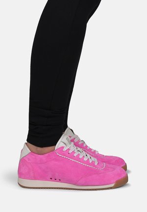 Osoba nosząca jasnoróżowe zamszowe trampki z białymi sznurowadłami i beżowymi podeszwami, w połączeniu z czarnymi dopasowanymi spodniami.