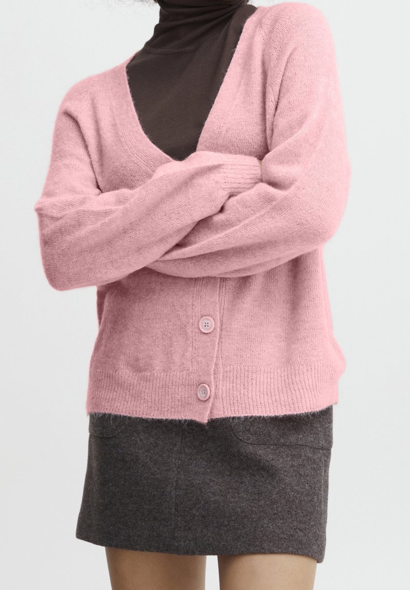 Rosa stickad kofta med V-hals och långa ärmar, prydd med två rosa knappar. Bärs över en brun polo och en grå kjol.