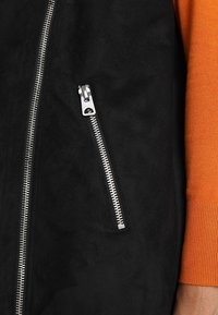 Svart mocka plagg med en silverfärgad dragkedjedetalj på vänster sida, kombinerat med en orange stickad ärm. Slät textur och modern design.