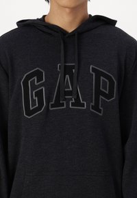 Czarna bluza z kapturem z wyraźnym logo "GAP" w ciemnoszarym kolorze, z regulowanym kapturem na sznurki i kieszenią z przodu, wykonana z miękkiego materiału.