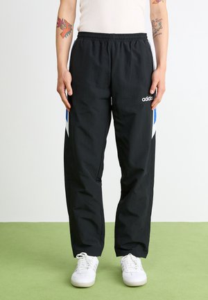 Osoba ubrana w czarne spodnie dresowe Adidas z niebiesko-białymi paskami po bokach, białe trampki i biały top bez rękawów, stojąca na zielonej podłodze.