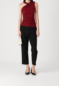 Burgundy sleeveless top with a knotted design, paired with black wide-leg trousers. Model holds a small white handbag and wears black heels.