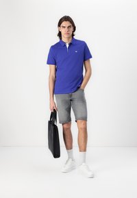 Mørkeblå polo med hvid krave, parret med grå denimshorts. Modellen holder en sort taske og har hvide sneakers på med grå sokker.