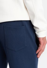 Marinblå byxor i en texturerad tygkvalitet, med en enda bakficka och kontrasterande sömmar. Midjan är delvis synlig.