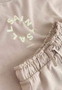 Ružová mikina a zodpovedajúce nohavice z mäkkého materiálu; obsahuje biely kruhový text "SAINT" a "LONDON"; elastický pás s detailom na šnúrku.