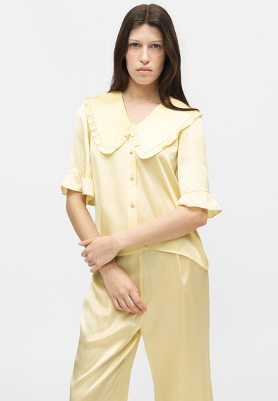 Jeune femme portant un ensemble de pyjama en satin jaune pâle avec un grand col à volants et un haut boutonné, debout devant un fond blanc uni.