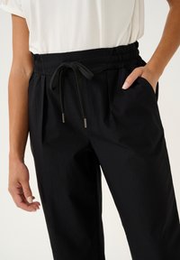 Pantaloni neri con vita elastica e cordoncino. Presentano due tasche laterali e una texture liscia con pieghe per un tocco di dettaglio in più.
